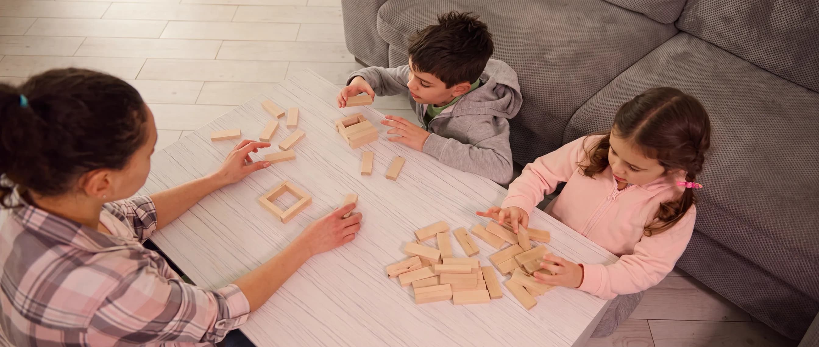 A parent and two young children playing together with wooden building blocks at a table at home.