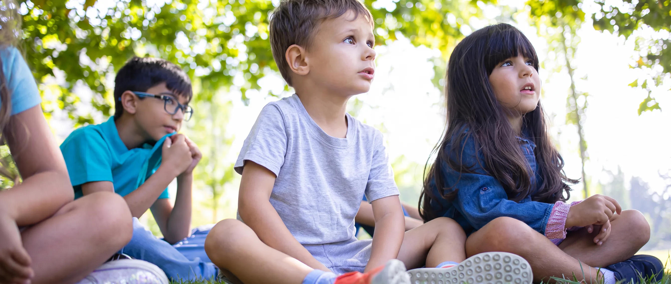 A group of young children sitting together outdoors on the grass, engaged and looking in the same direction.