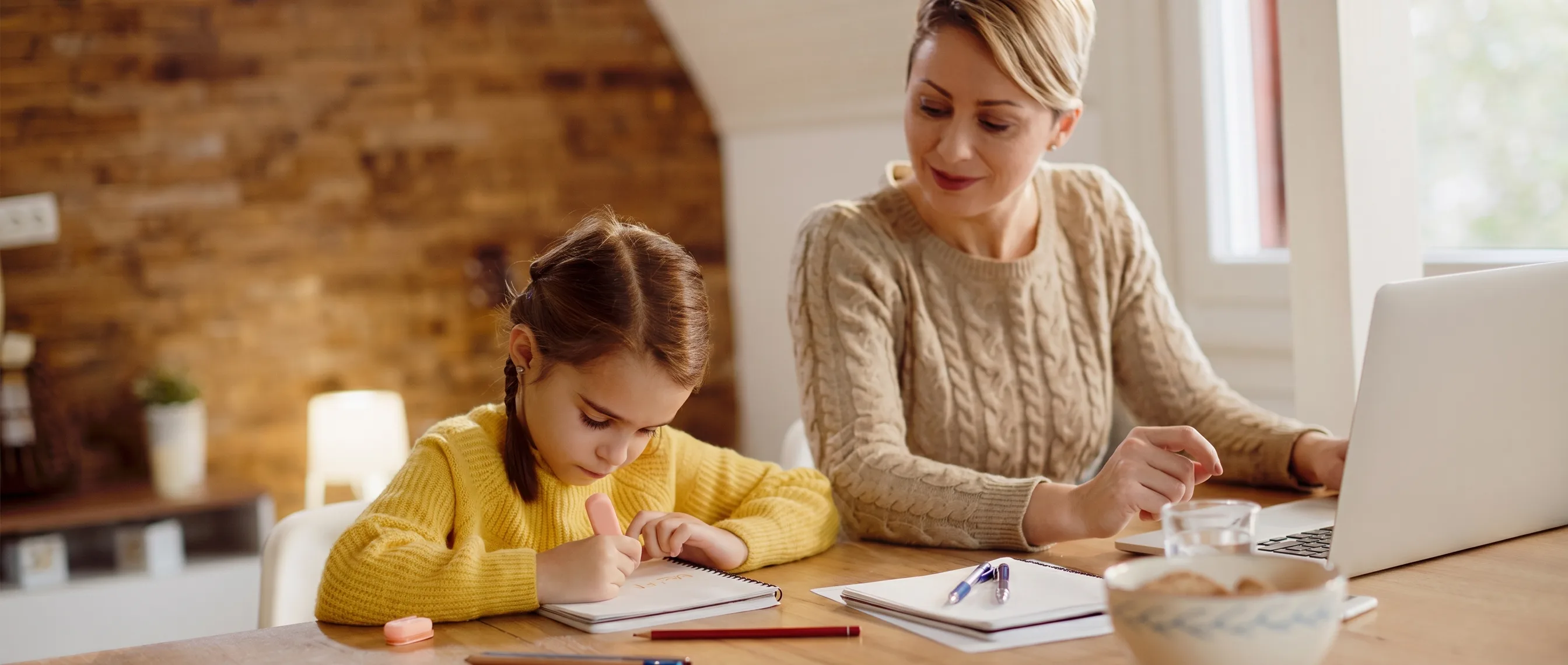 A parent working at a laptop with notebooks and paperwork at the kitchen table, while their young child draws in a notepad beside them.