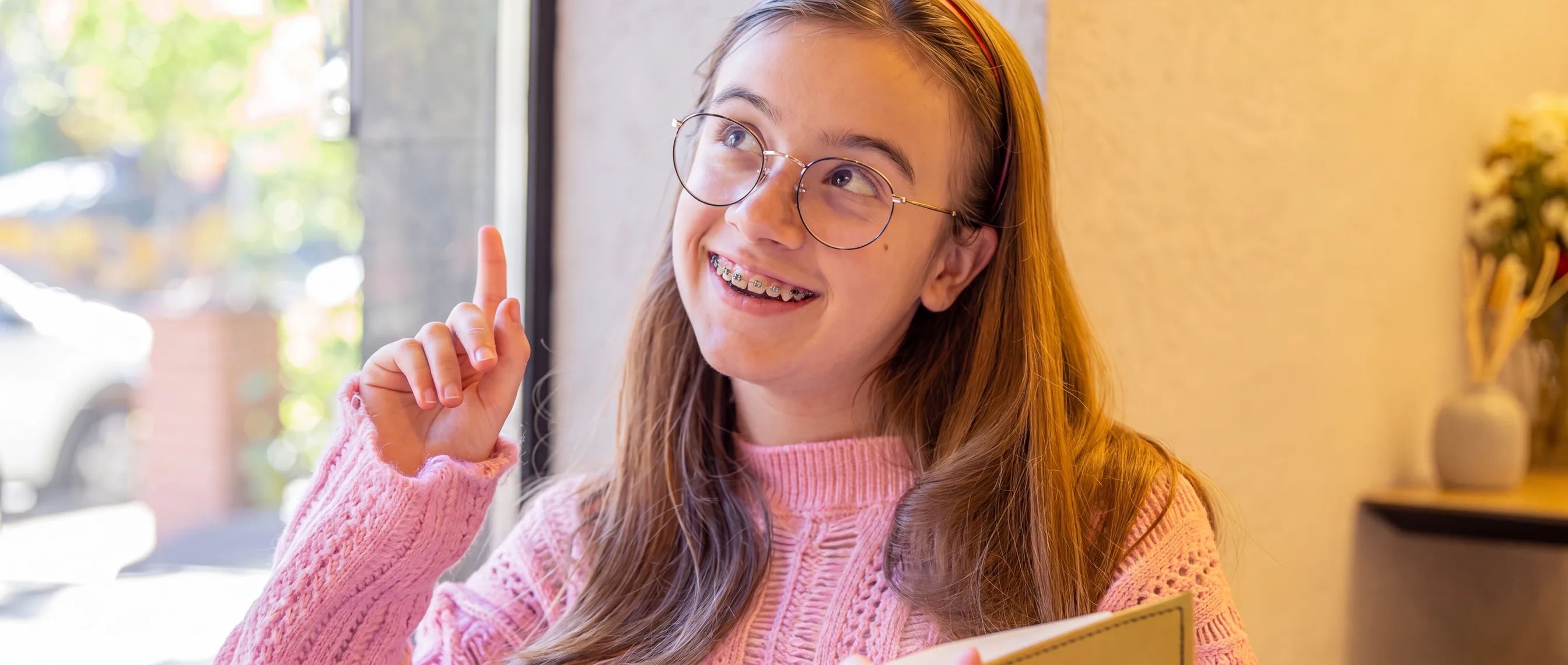 A smiling teenage girl holding an open book, looking up as an idea occurs to her.